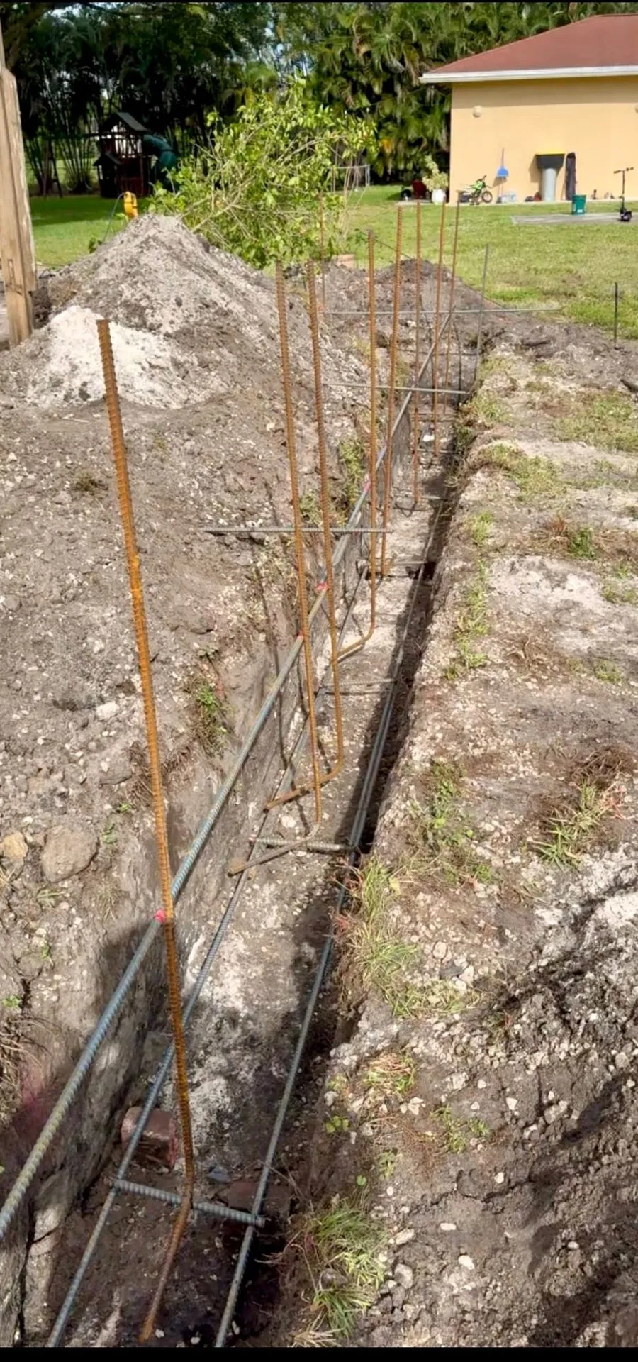 Reinforced concrete footing trench with rebar stubs along a backyard