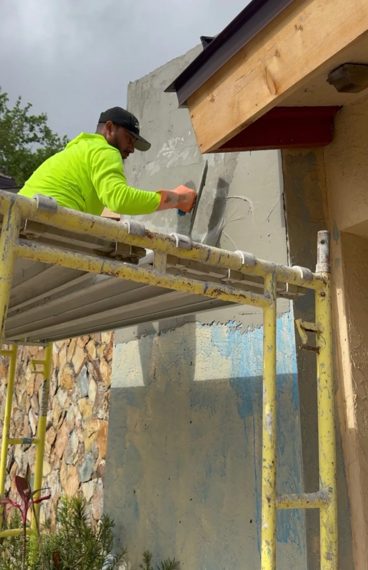 Contractor applying stucco from scaffolding beside a stone veneer wall